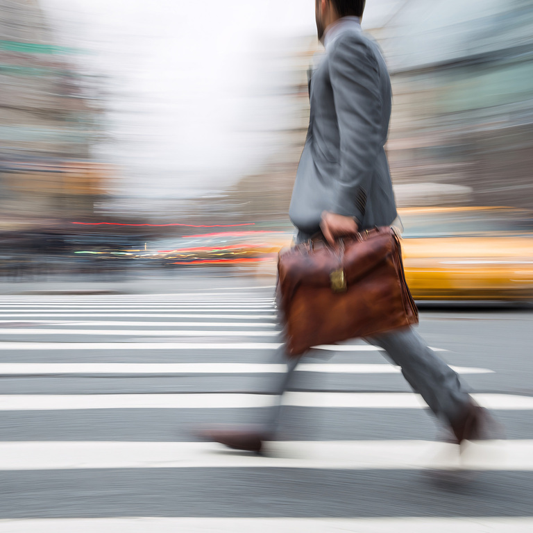 A blurred businessman hurrying through a busy city crosswalk, capturing the pressure and pace of modern work.