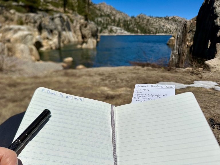 A hand and pen journaling with the scenery of a beautiful alpine lake in the background