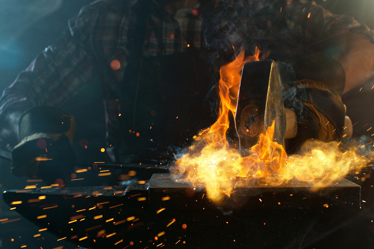 A smithy works a hot, flaming piece of steel with a forge hammer